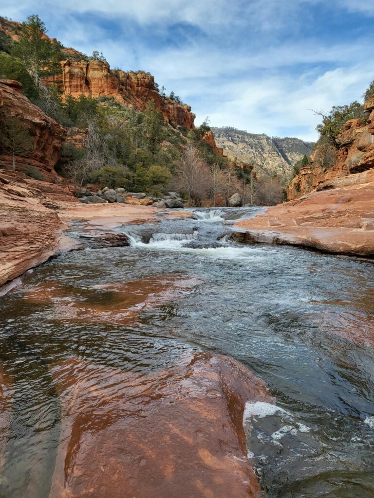 A tranquil stream flows through red rock canyons under a clear sky in Sedona, Arizona.