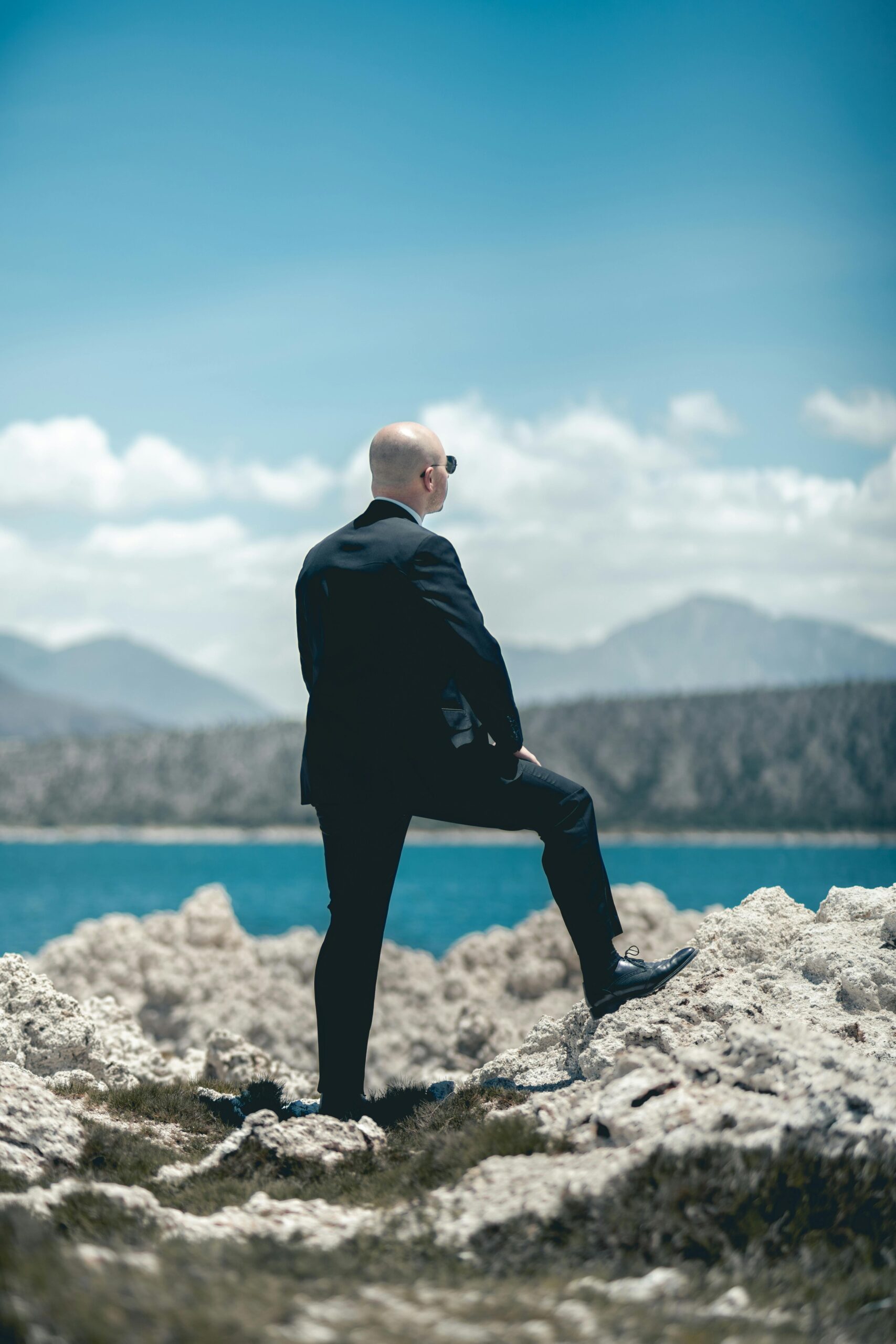 Elegantly dressed bald man in a suit stands on rocks, gazing at a scenic lake and mountains in Puebla, Mexico.